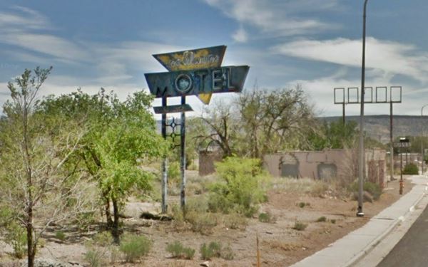 color view neon sign of a motel, 1950s, triangular blue and yellow, faded; set in an overgrown lot. Route 66 to the right