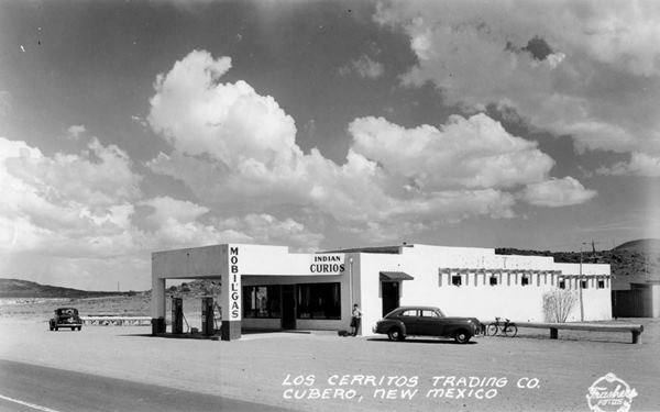 black and white 1940s picture Mobil gas station, flat canopy, two cars, 2 gas pumps, person by office, Trading Post written on Postcard- Indian curios written on building, US66 in front, hills beyond