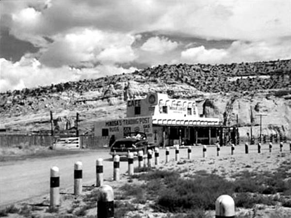 black and white still from the movie, 1951, car driving up to 2 story adobe style pueblo bldg, a trading post, hill beyond, gas pumps