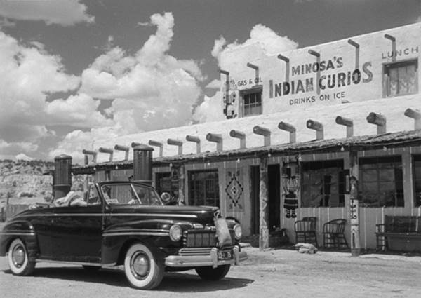 black and white still from the movie, 1951, convertible car parked by 2 story Pueblo style trading post, vigas on roof, gas pumps