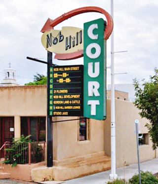 image showing neon sign with yellow shape on top with words NOB HILL, encircled by a red arrow, and a vertical rectangle painted green with the words COURT in white, motel units and sidewalk.
