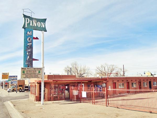steel fence faces Route 66, classic Neon sign, faded blue with white vertical word MOTEL, atop, green background word PINON in white. Pueblo style single floor ochre color building to the right