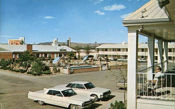 1960s postcard of the inner courtyard of the Ramada, surrounded by two story gable roof units, a pool. Cars, man on room verandah