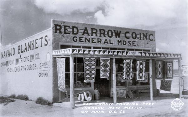 single story bos shaped stucco flat roof building parapet reads RED ARROW CO. INC. GENERAL MDSE. Rugs hang from awning held up by posts, NAVAJO BLANKETS CAMP GROUND INDIAN JEWELRY & CURIOS painted on side wall, seen from dirt street