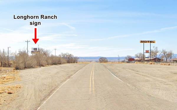 US 66 heading away, hills in the distance, motel on the right with neon signs, rusty sign among some dry trees to the left