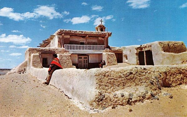 adobe church, low wall around atrium, choir balcony, cross atop a stubby bell tower, person sitting on wall, color 1950s