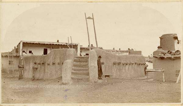 sepia, 1880. Adolph Bandelier standing outside of a round adobe kiva, ladders and adobe pueblo behind