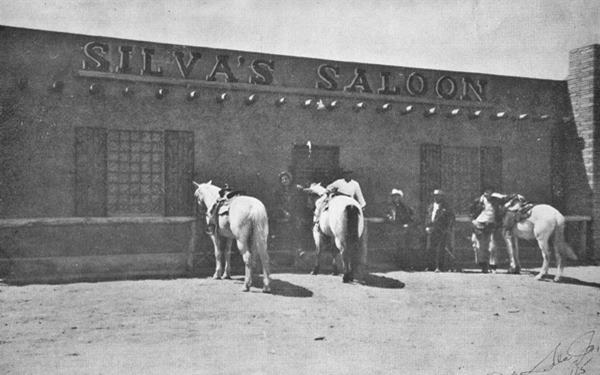 black and white, adobe flat single story bar SILVA’S SALOON written on parapet, five men and 3 horses in front.