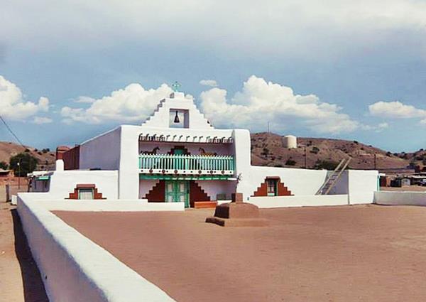 color white adobe church, with balcony, stepped parapet above it with bell. Vigas on front wall, low wall encloses atrium, hills beyond