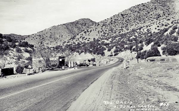 black and white postcard from 1940s, US 66 to the right, curves left in the distance. Hills with trees.  Box shaped stone building, with black car to the left