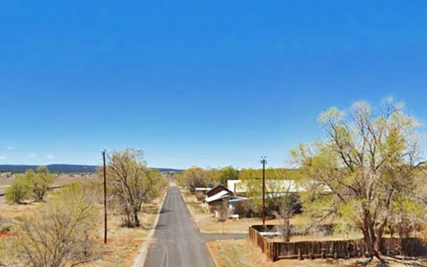 street heads west, lined by some trees, red dirt in the yards, some houses, hills-mesas in the distance it is the old 1926 to 1937 Route 66 in Thoreau, New Mexico