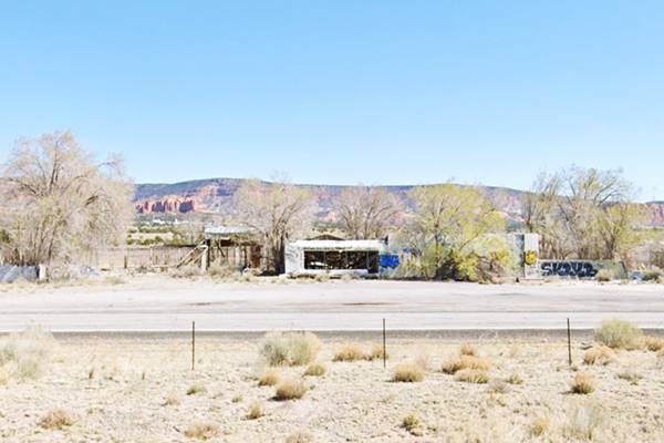 color photo. long single story building vacant and in ruins, trees around it, red cliff on mesa in the distance, US 66 runs in front of it