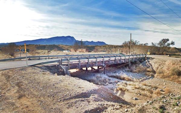 color: Bridge seen sideways, timber piers, gravel dry creek, highway runs across, mountains in the distance