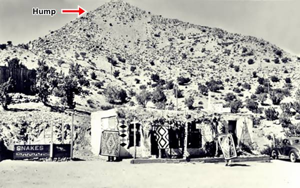 black and white picture 1930s cars right, trading post middle, SNAKES sign left, two men hold up native woven carpets, hump-shaped hill behind