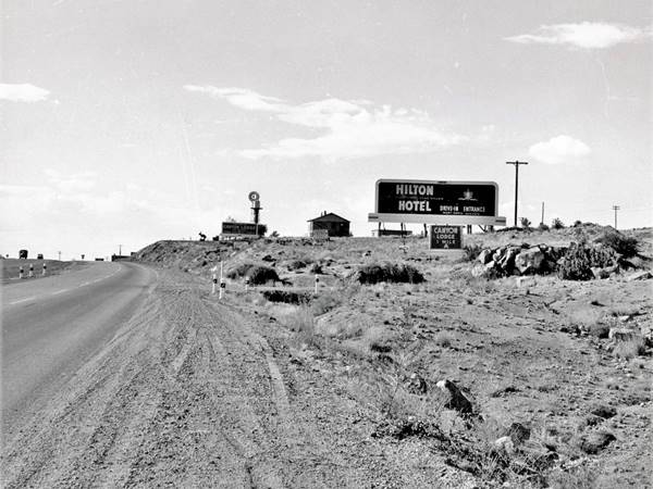 black and white photo looking west taken on westbound lanes shoulder, truck on left is on the eastbound lanes. Ahead signs of motels, hotels, a windmill and a gable roof building.