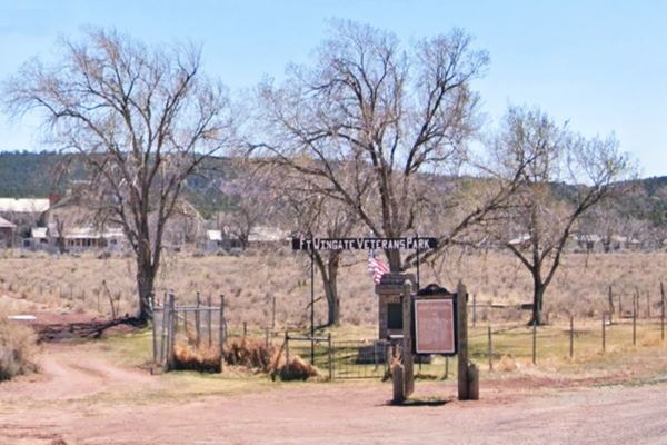 Small park at a fork, sign reads VETERANS PARK, stone monument, Fort Wingate Marker, lawn, wire fence, and trees. Hills in the distance