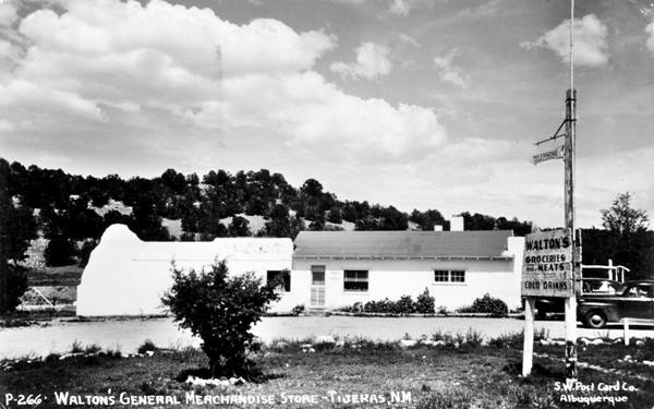 black and white postcard from 1948, US 66 runs across the image. Behind a building, sign reads Walton’s Store, hills with trees, and 1940s car