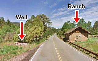 color; highway runs ahead in the middle, to the right gable roof brown ranch building. Left: rock circle, a well, trees line the highway