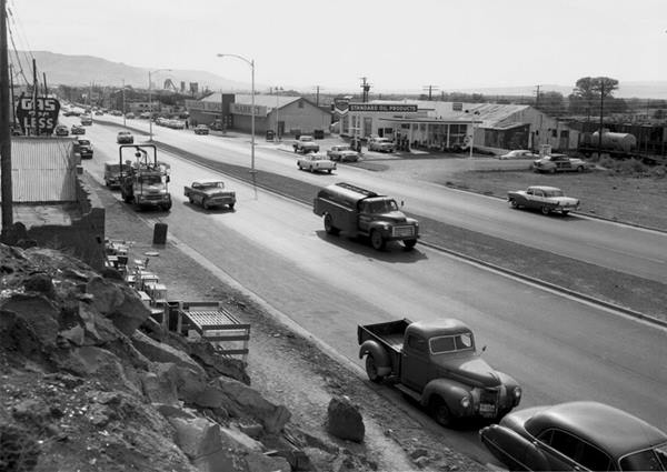 1950s black and white: highway, to the right a Chevron station, gable roof building, a trading post. To the left, garage, gas station and beyond the Yucca Hotel, cars and truck on US66