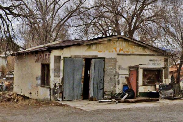 White Arrow Garage San Fidel US66 gable zinc roof, wood frame building with stucco on walls. Words on upper part say WHITE ARROW GARAGE, gravel drive, trees, color picture