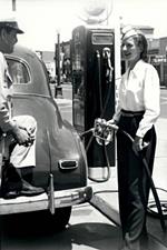 1940s woman pump attendant Firestone Service Store black and white photo, lady pumping gas into a vintage car, man with foot on bumper talking to her