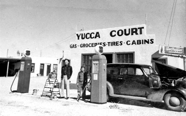 1940s black and white photo, gas pumps, man and child, building with words GAS. GROCERIES. TIRES. CABINS, on office