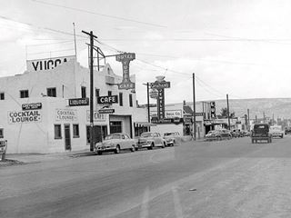 1950s view of Route 66 with cars in the foreground and buildings along it: Yucca Hotel, stores, neon signs and people. Black and White