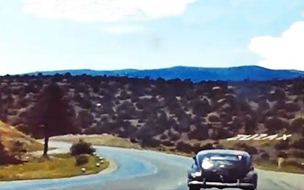 color still from a video taken in 1958: four lane highway, car ahead, hilly terrain, word ZUZAX written in white stones on the right. Tree to the left