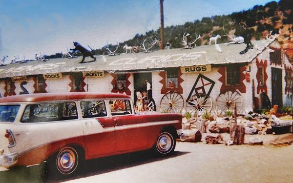 color picture c.1950s of a gable roof store, antlers on roof, advertising on roof, signs of rugs, jackets, rocks. Station wagon and hill behind