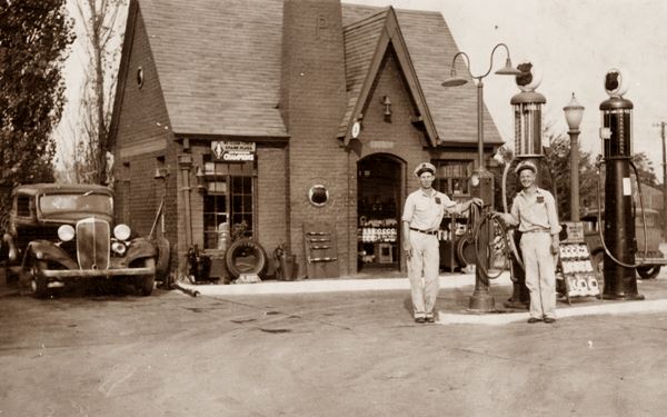 Ancient black and white photo, two men, gas pumps, cottage-like building and 1930s car