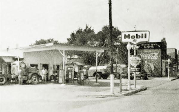 black and white picture of a Mobil gas station on a corner, people, and a large Coca Cola sign on the adjacent builiding
