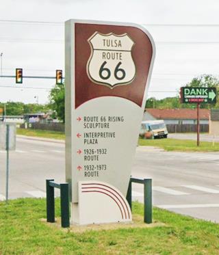 US 66 marker panel sign in brown with Route 66 shield and text with arrows showing nearby sights on grassy median in Street