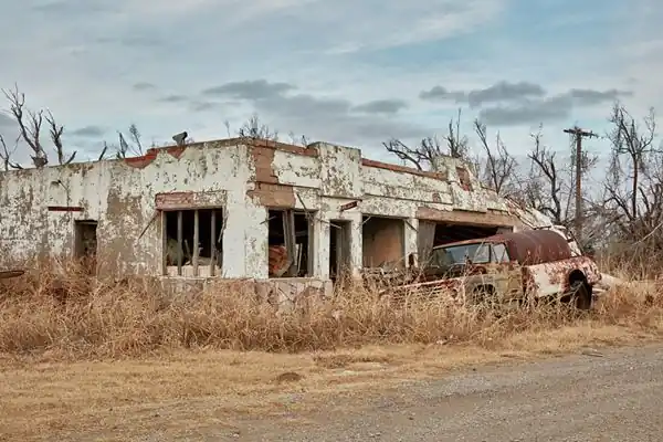 ruined one story cafe among brown dry overgrown grass, with rusting truck in front of it