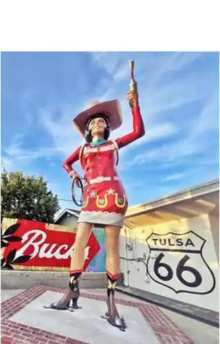 20 ft. tall woman statue in boots, with red dress with white details, turquoise scarf, cowboy hat, space gun up in the air in right hand, US66 shield behind