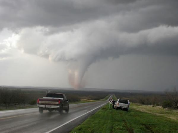 Tornado TX highway heads towards dark cloudy sky, tornado to the left in the distance. Pick up drives by on wet highway, another parked on grassy shoulder